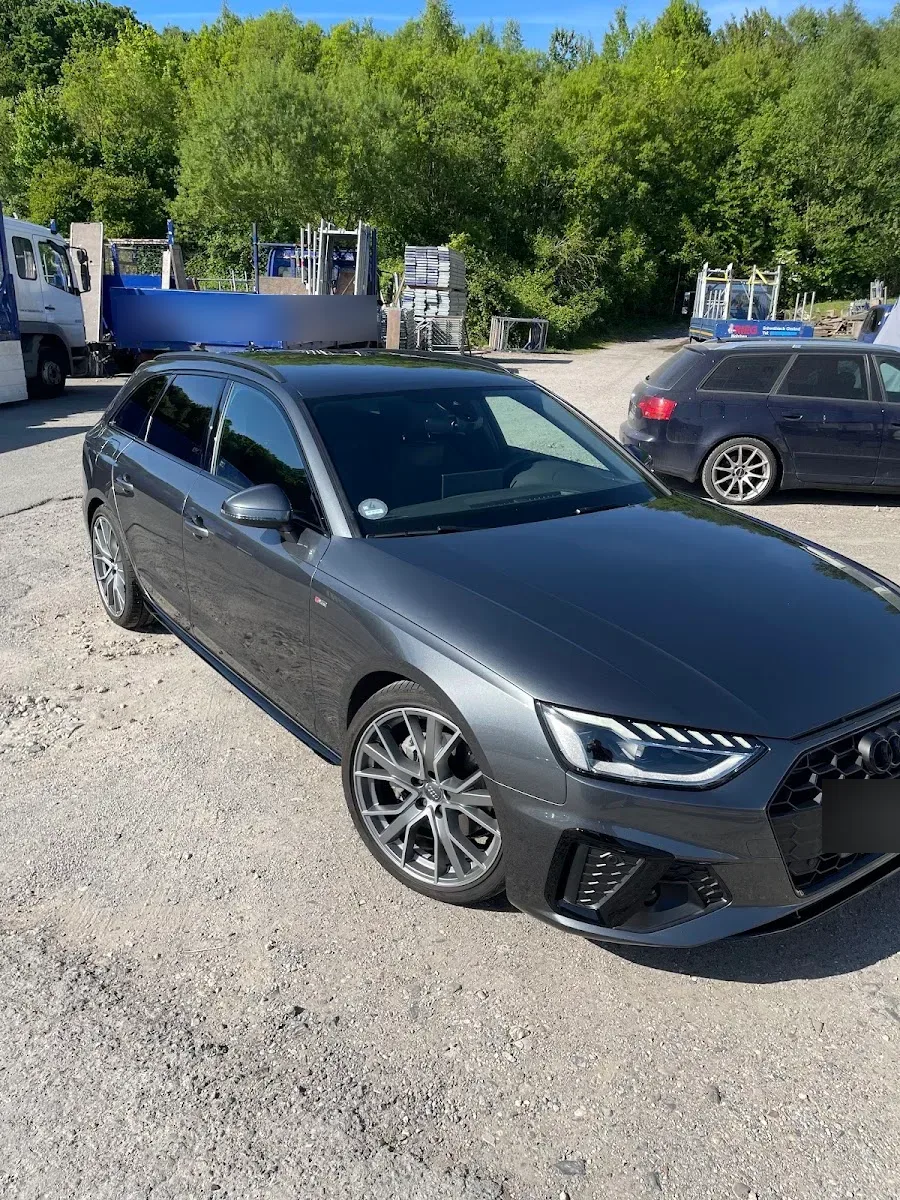 Grauer Audi A4 Avant S-Line mit glaenzendem Lack steht bei Sonne auf einem Schotterplatz vor Baustellenfahrzeugen.