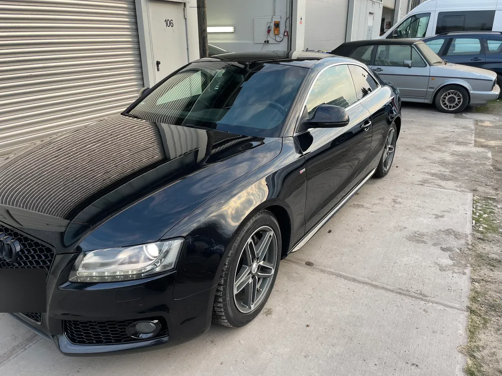 Schwarzes Audi A5 Coupe S-Line vor dem Hallentor, Lack spiegelt Tor und Himmel, Scheinwerfer eingeschaltet.