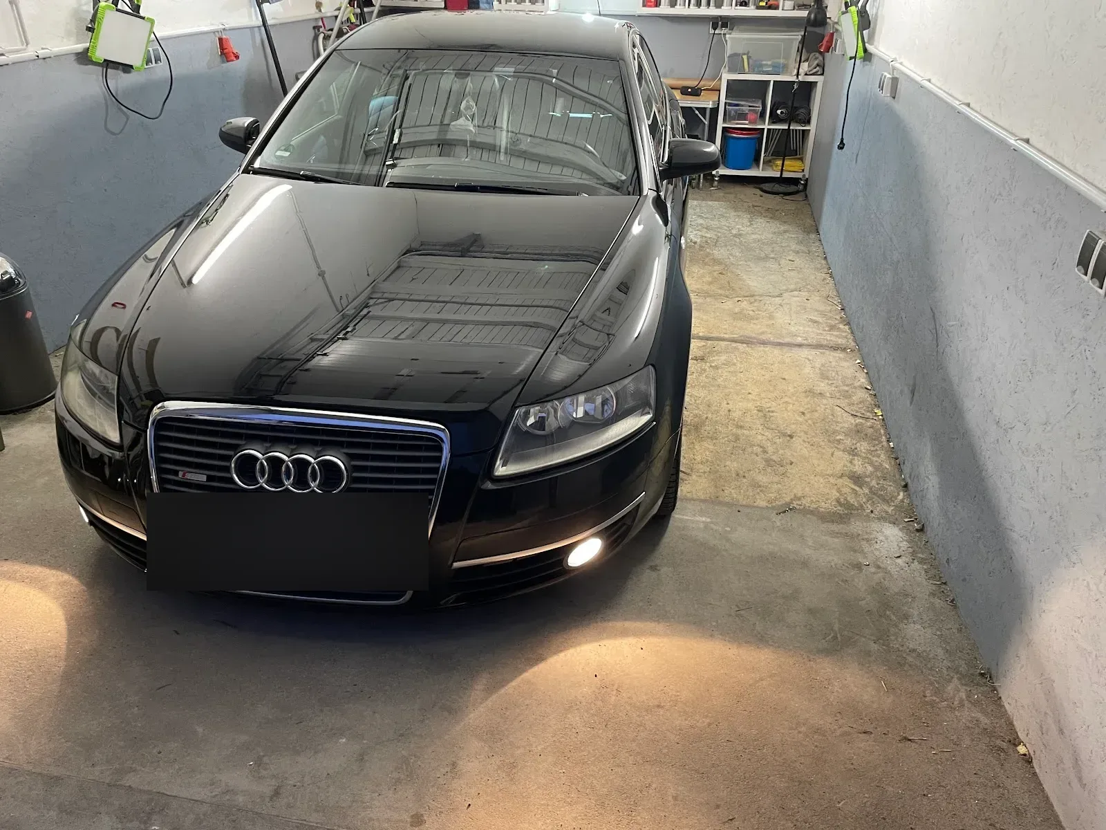 Schwarzer Audi A6 Avant steht frontal in der Aufbereitungshalle, Lack glaenzt stark, Tagfahrlicht an.