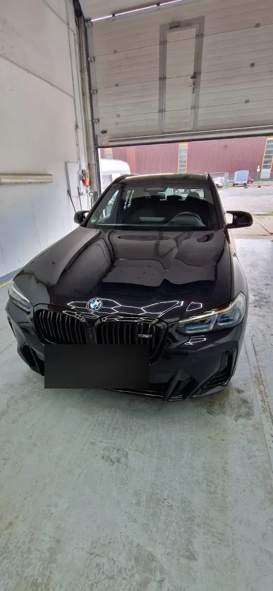Schwarzer BMW X3 M-Paket steht hochglaenzend poliert im Hallentor, die Deckenstruktur spiegelt sich auf der Motorhaube.