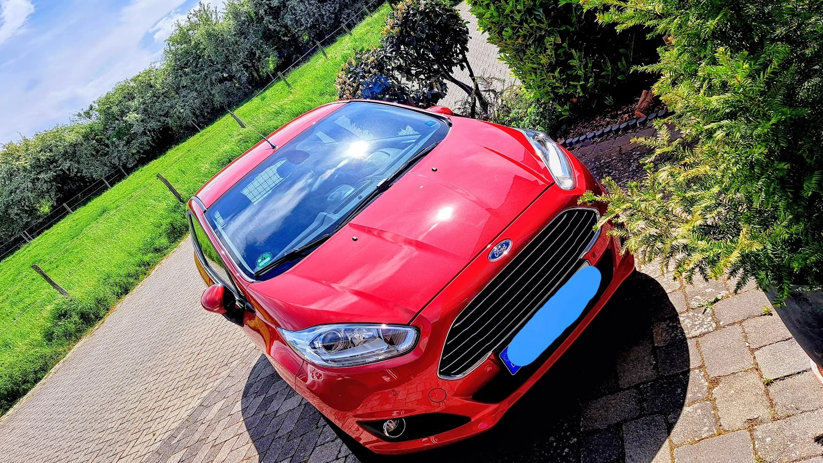 Roter Ford Fiesta von schraeg oben auf einer Pflastereinfahrt, der Lack glaenzt stark in der Sonne, das Kennzeichen ist unkenntlich gemacht.
