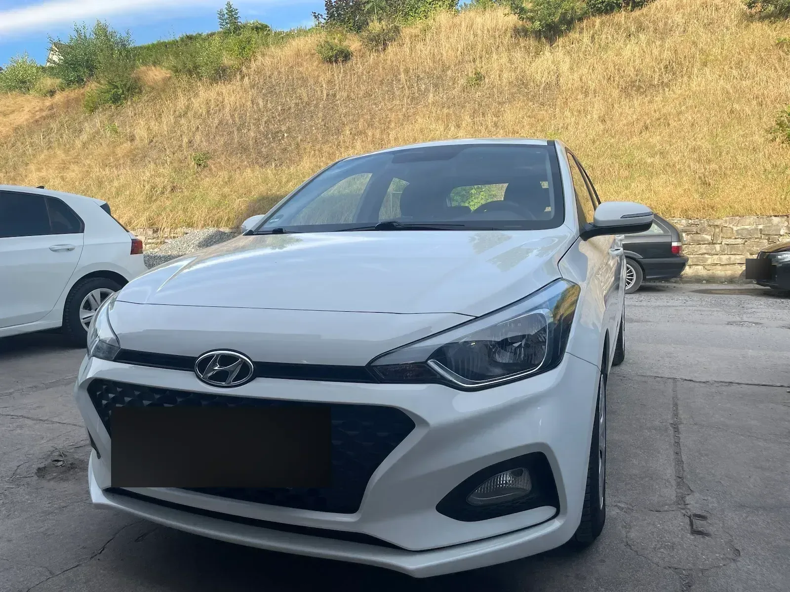 Weisser Hyundai i20 frontal-schraeg auf dem Hof, Lack sauber und glaenzend in der Abendsonne.