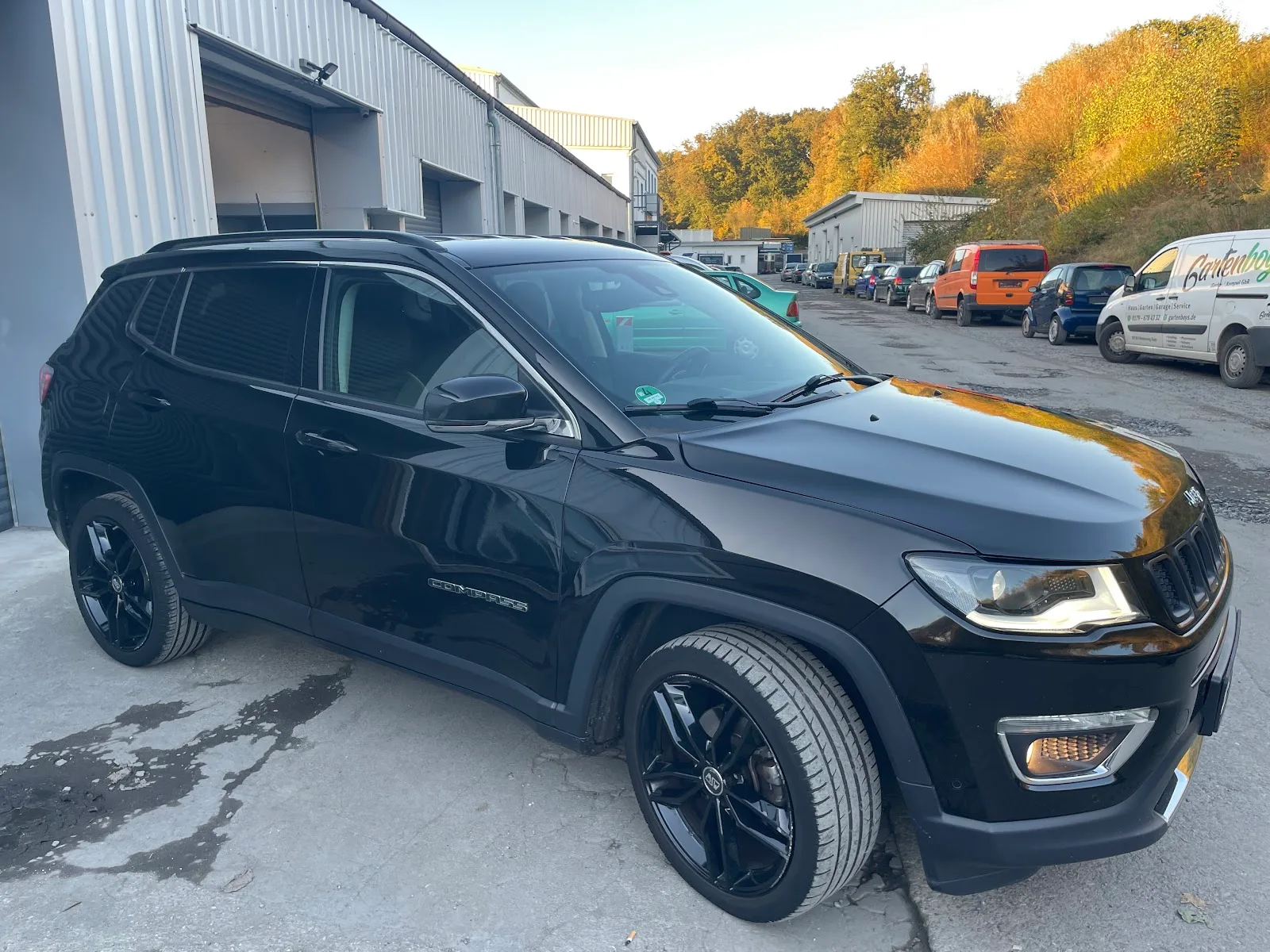 Schwarzer Jeep Compass in Dreiviertelansicht vor der Halle, Lack spiegelt die Abendsonne, schwarze Felgen.