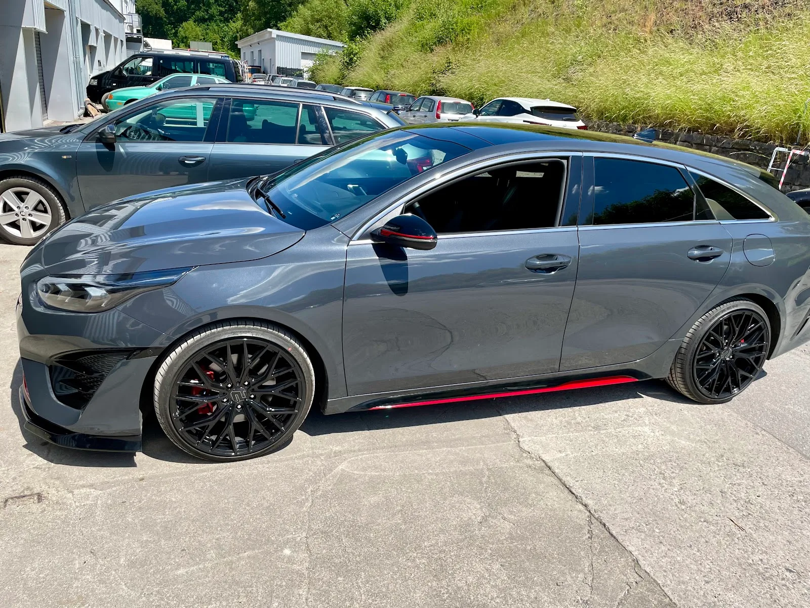 Grauer Kia ProCeed GT Shooting Brake in Seitenansicht auf dem Hof, Lack spiegelt stark, schwarze Felgen und rote Zierleiste.