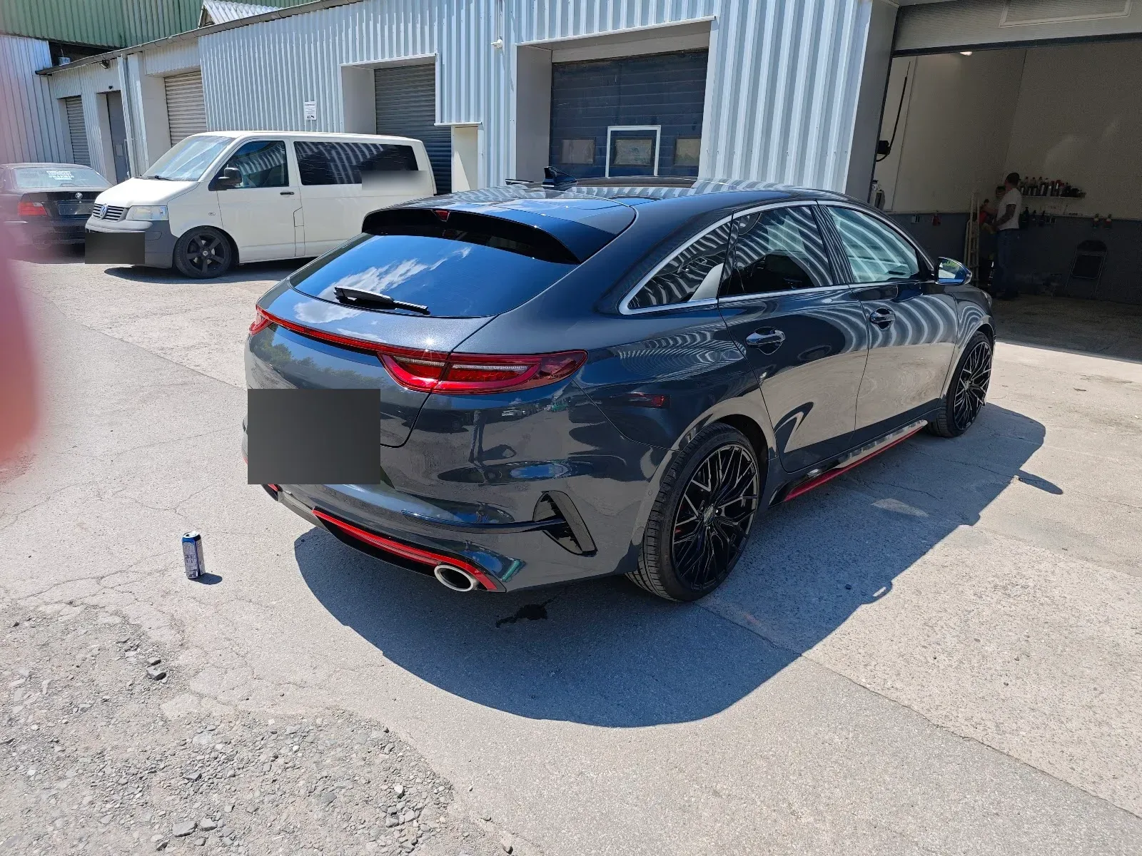 Dunkelgrauer Kia ProCeed GT von hinten in der Sonne mit glaenzendem Lack und schwarzen Felgen, links ein roter Lichtschleier vom Finger vor der Linse.