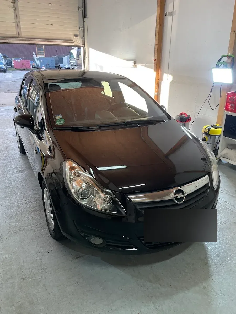 Dunkelbrauner Opel Corsa D steht im Hallentor, Lack und Scheinwerfer sauber, Sauger im Hintergrund.