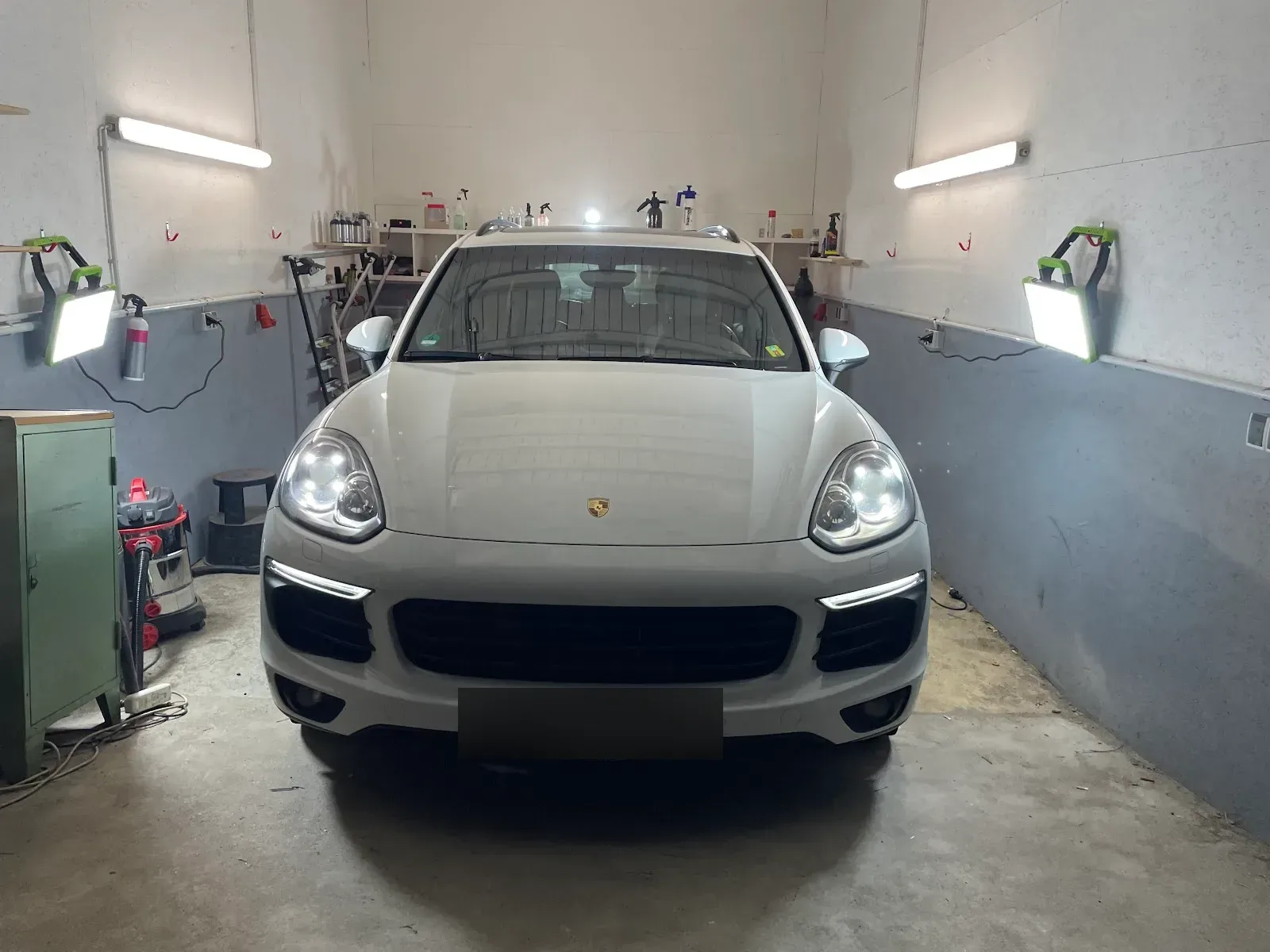Weisser Porsche Cayenne mit eingeschaltetem Licht in der ausgeleuchteten Aufbereitungsbox, Arbeitsleuchten und Pflegemittel-Regale im Hintergrund.