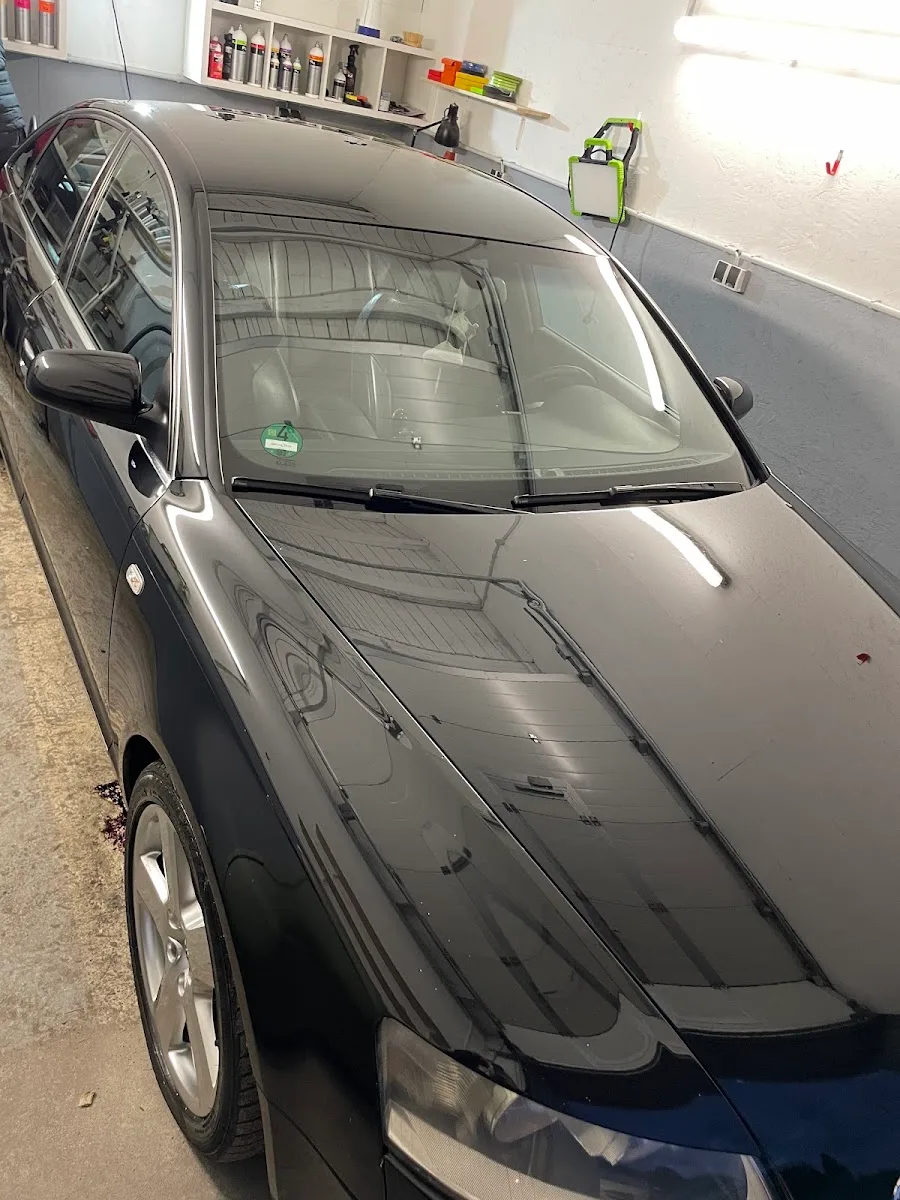 Schwarzer Audi A6 in der Halle, Motorhaube und Dach spiegeln die Hallenbeleuchtung, im Hintergrund Regal mit Pflegemitteln.