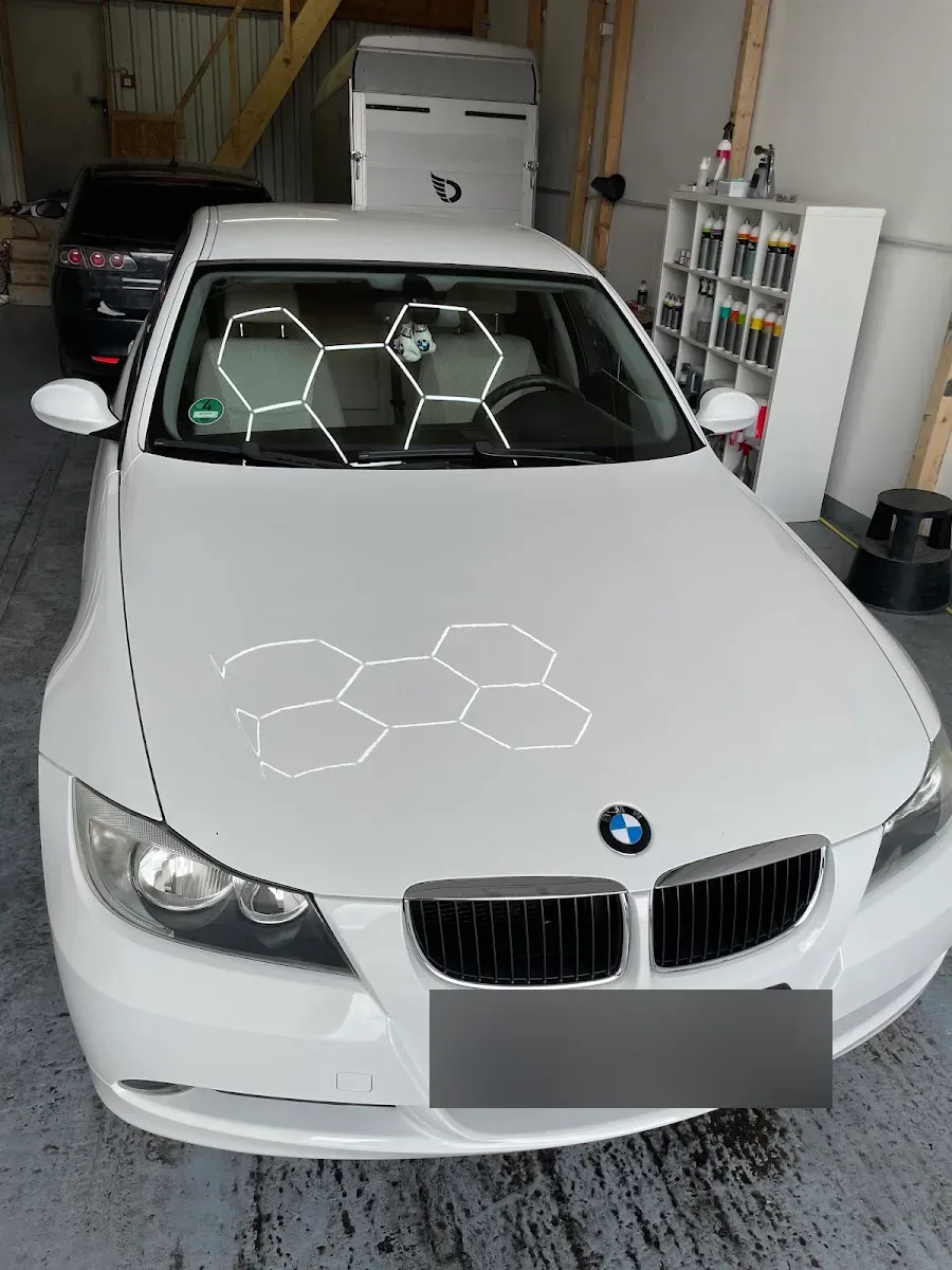 Weisser BMW 3er E90 in der Halle unter Hexagon-Lichtern, deren Reflexion die makellose Motorhaube zeigt.