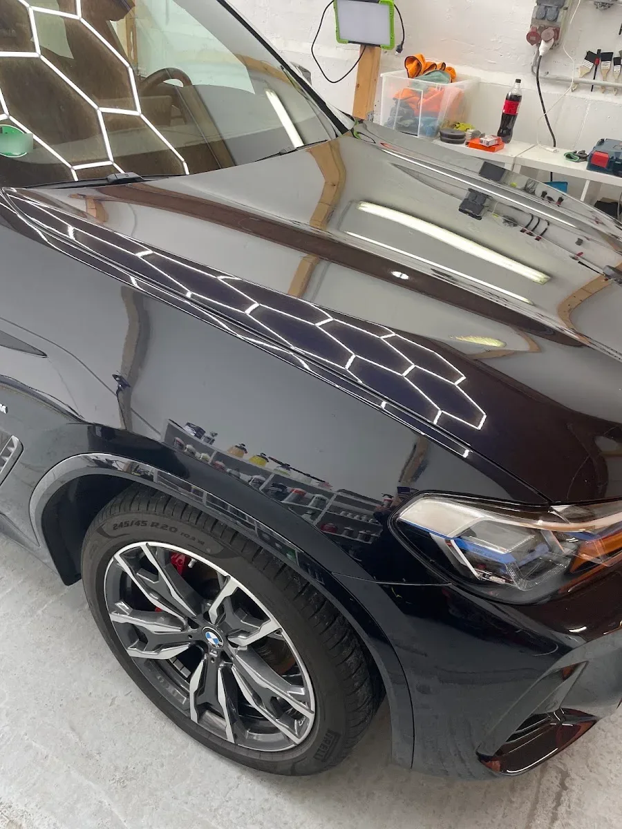 Nahaufnahme von Motorhaube, Kotfluegel und 20-Zoll-Felge eines dunklen BMW, Hexagon-Leuchten spiegeln sich messerscharf im Lack.