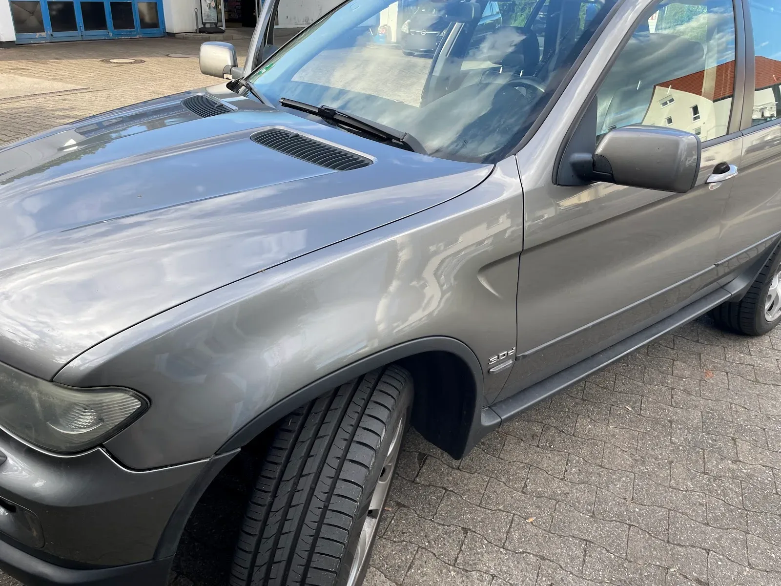 Nahaufnahme der polierten Motorhaube und Flanke eines grauen BMW X5 E53, Reflexionen zeigen den aufbereiteten Lack.