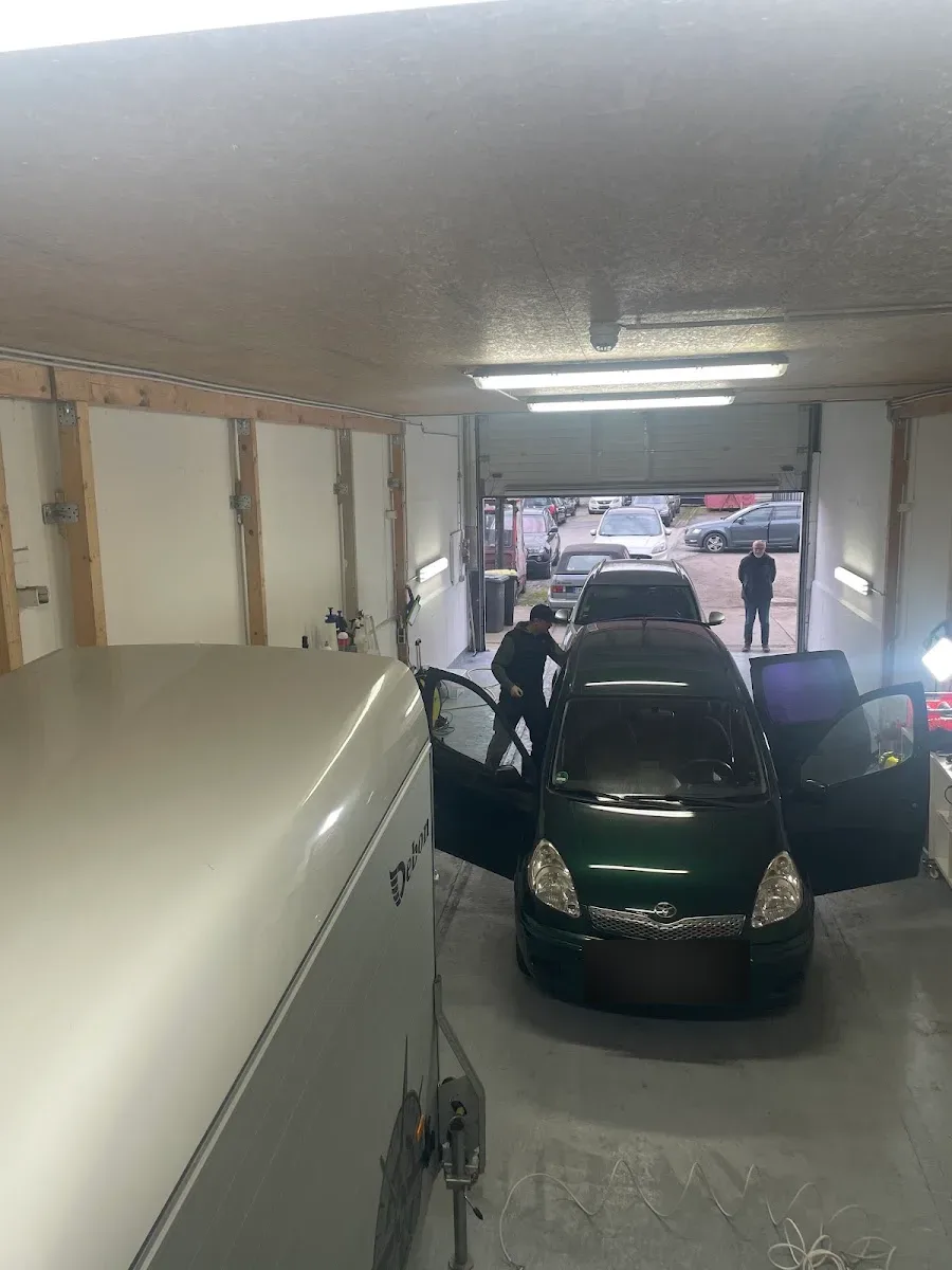 Blick in die Aufbereitungshalle: gruener Toyota Yaris mit offenen Tueren wird bearbeitet, links ein Wohnwagen, im Hintergrund das offene Hallentor.
