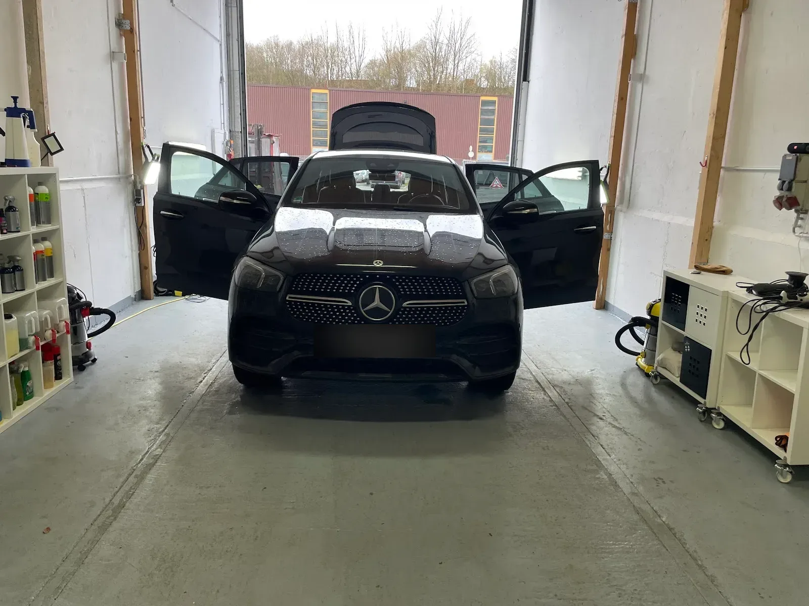Schwarzer Mercedes GLE Coupe mit geoeffneten Tueren und Heckklappe in der hellen Aufbereitungshalle, Pflegemittel-Regal und Sauger im Bild.