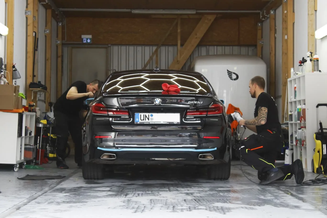Zwei Mitarbeiter polieren einen schwarzen BMW 5er in der Aufbereitungshalle, einer mit Poliermaschine am Heck, Wabenlicht spiegelt sich im Lack.