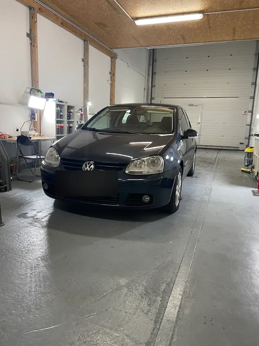 Dunkelblauer VW Golf 5 steht in der beleuchteten Aufbereitungshalle, Frontansicht mit Arbeitsplatz und Regal im Hintergrund.
