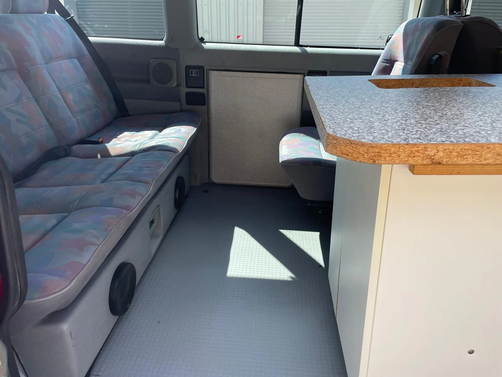 Innenraum eines VW-Bus-Campers mit gereinigter gemusterter Sitzbank, Kuechenblock und sauberem Riffelboden.